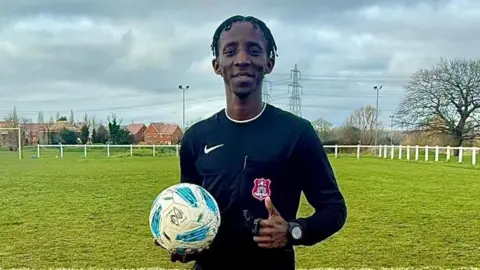 George King/BBC A young man wearing a black referee kit while holding a football in one hand. He is standing on a grass football pitch and looking into the camera.
