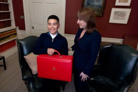 A young boy is pictured with Chancellor Rachel Reeves. He is holding the famous "red box" which ministerial papers are transported in.