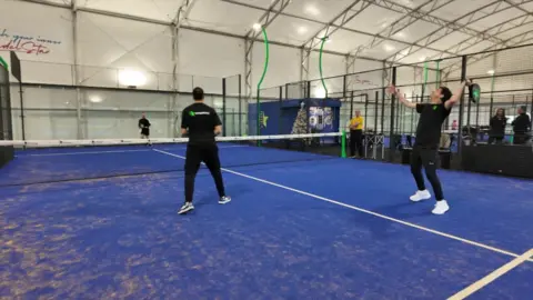 Three padel players dressed in black on a blue court, similar to an indoor tennis court with a large metal-framed walls and roof  