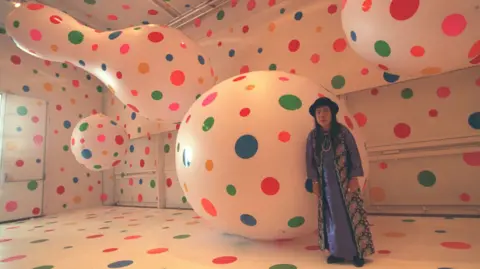 Getty Images Yayoi Kusama standing among giant white shapes with multi-coloured polka dots