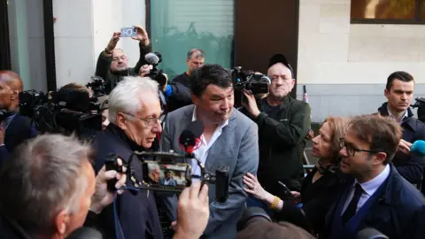 PA Media Graham Linehan at the centre of a small group of people outside court, some pointing cameras at him and speaking to him