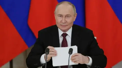 EPA Vladimir Putin. The Russian president is seated in front of a Russian flag and behind two microphones and is holding a piece of paper as he makes an address. He is wearing a dark suit, white shirt and red tie