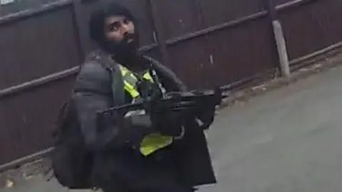 Body worn camera still of Hari Mann on a street, dressed in black with a hi vis vest and holding what looks like a  short black machine gun