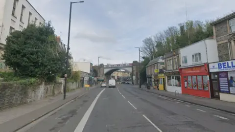 Railway bridge passing over Cheltenham Road in Bristol. The road is lined with houses on the left and shops on the right.