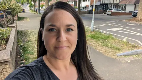 Sarah A woman stands on a residential street. She is wearing a grey T-shirt and she has straight black hair.