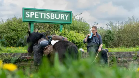 sesmlphotography Roger and Scarlet having a break from their trek near a road sign for Spetisbury. Roger is drinking from a water bottle. It is cloudy.