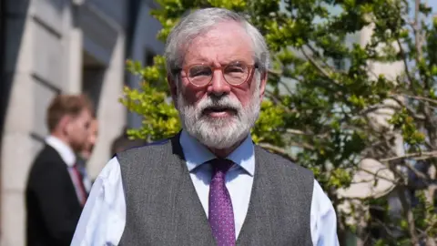 Gerry Adams, wearing a blue shirt, purple tie and grey waistcoat. He has grey hair and a grey beard and wears brown-rimmed glasses. The court is behind hin, with trees outside.