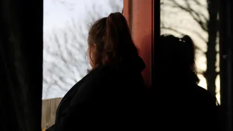 An image of a woman, whose back is to the camera and who is in partial shadow, looking out of a window of a house. She cannot be identified from the photograph. 