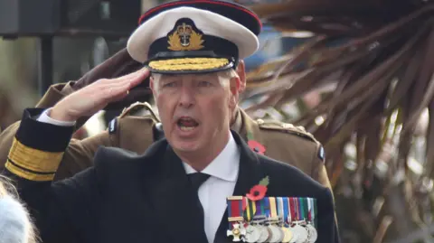 Jonathan Carley dressed as an admiral gives a salute. He wears a white hat, navy jacket, white shirt and tie. On his chest are a series of medals hung by colourful ribbons. 