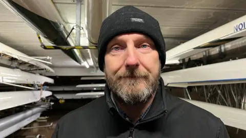 Stewart Plant is standing in a boat house with white boats stacked around him. He has grey and brown facial hair and is wearing a black beanie and black jacket.