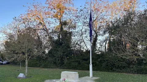 A purple flag at half-mast at a memorial, on a grass lawn, with trees in the background. There is blue sky above.
