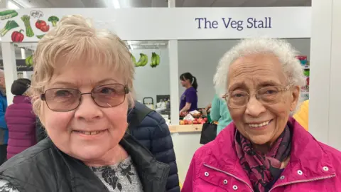Jean and Joan, two women with short hair, smile into the camera as they stand in front of a fruit and veg stall. Jean, on the left of the image, is wearing tinted glasses, a black and grey top and a black gilet. Joan, on the right of the image, also has glasses and is wearing a bright pink coat.