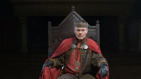 Head is dressed as a medieval king, and sits on a throne, for a press picture 