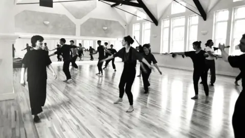 Circomedia An old black and white image of students at Circomedia in a dance hall being trained. They look like they are moving with their arms outstretched and they're all mainly wearing black.