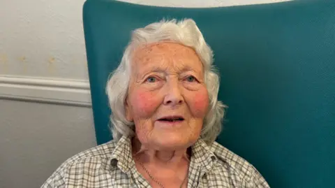 Sally wearing a check shirt and has grey hair. She is looking at the camera