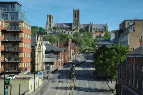 Graham Halliday A photo showing a dual-carriageway road leading towards a large cathedral on a hill under a blue sky. Large buildings line either side of the road, while green trees are also dotted along the route.