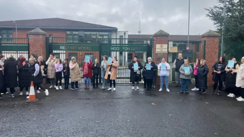 A group of parents and guardians outside the school gates holding blue signs.