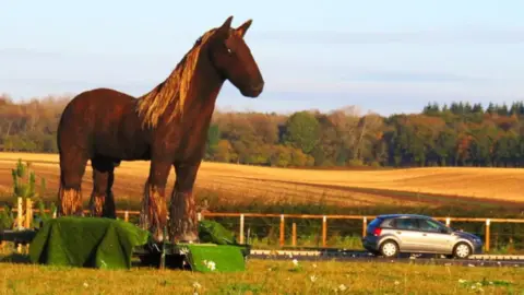 Damian O'Connor A sculpture of a horse made from steel, wood and reeds displayed by a road in Norfolk. On the right of the picture is a silver car.