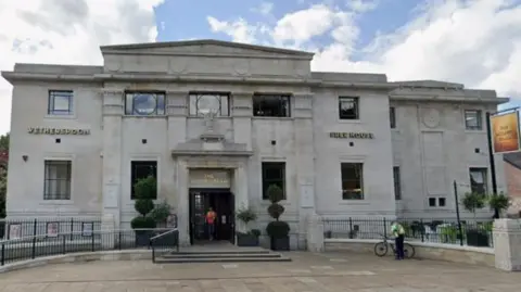 The image shows the front of a large stone building positioned along a paved pedestrian area, with a clear, bright sky in the background and scattered clouds.
The main entrance is framed by two tall rectangular windows and large potted plants. A sign above the door reads "The Golden Beam".
A person wearing a bright top is standing in the doorway, and another person with a bicycle is visible next to a railing on the right of the image.