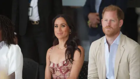 Getty Images Harry and Meghan sit next to each other smiling. Other people can be seen sitting beside them and standing behind them. 