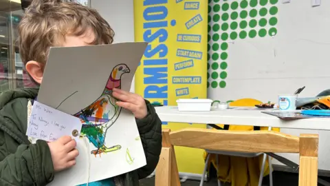 Permission 2 A young boy in a green coat covers his face with his design and a note to his nanny. A work table with art supplies and a wooden chair are in the background.