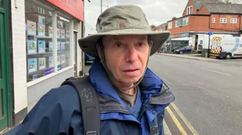 LDRS Dave Cooper is standing on the high street in Cheadle. Int the background is an estate agents. He is wearing a hat and waterproof coat along with a backpack.