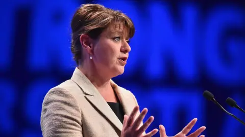 Getty Images Leanne Wood, with brown hair and wearing a cream suit, stands against a blue backdrop