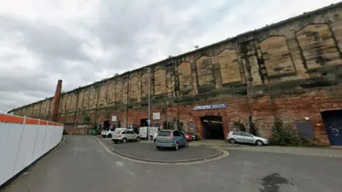 Google How the land where George Court will be built currently looks. A single carriageway turns to the left and runs beside the stone railway building. A number of cars are parked haphazardly on the pavement.