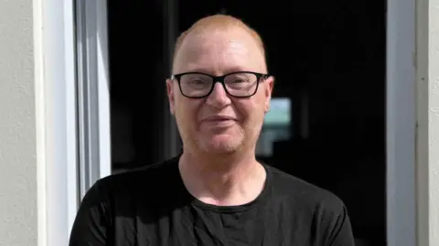 Dave Morris pictured wearing a black T-shirt with New York written on in pink. He has very short ginger hair and stubble. He is wearing black glasses and stood outside, in front of a door.