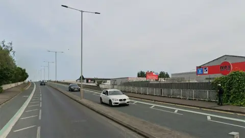 Hessle Road flyover with cars driving over it and a man walking down the right hand pavement. It has on-road cycle lanes and street lights along the central reservation. There are warehouses in the background.