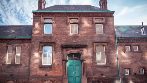 Friends of Carlisle Victorian & Turkish Baths The exterior of the Turkish baths building is made of orange and brown brick with a large green double door. There are regular rectangular windows with white frames set into the sides of the building. The sky above is bright blue between thin cloud cover.