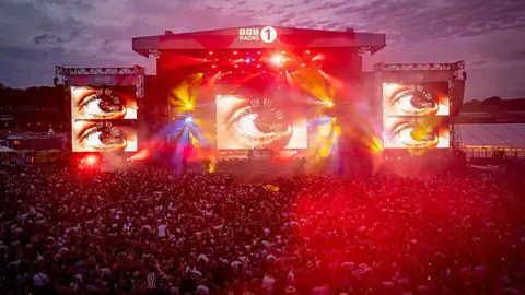 An ariel view of Fatboy Slim performing on the Big Weekend main stage. The screens show images of a big eyes. Bright red, yellow and blue lights shine across the crowd. 