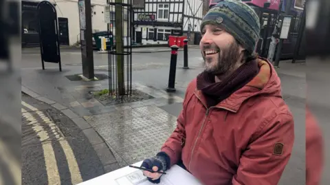 Artist Ric Liptrot smiling as he is sat drawing a shop front.