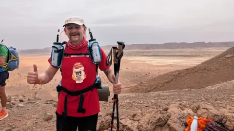 Lenny Worthing A man standing in a desert on the top of a sand dune wearing a red top and black trousers, as well as running vest with two water bottles. He is holding walking poles in his hand and holding his thumbs up. A vast desert is behind him.