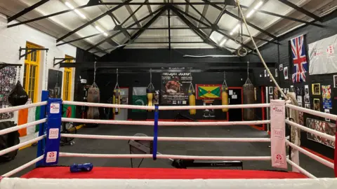 Alexander Smith A boxing ring in the foreground, and various punch bags around the wall of the sports club