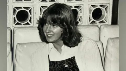 Lynne Darcy An old black and white picture of Lynne Darcy. She is around 14 years ols and has shoulder length hair which is styled and flicked back from her face. She is wearing a white cardigan over a white blouse and darker coloured top. 