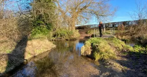 Action for the River Kennet (ARK) Woman standing on bank of small shallow river with grassy banks along its sides.