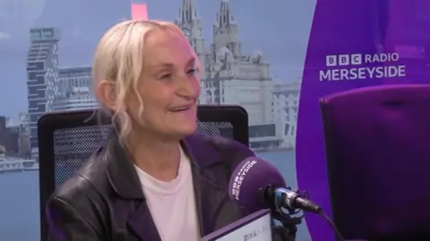 Mandy Jamieson has blonde hair and is wearing a white t-shirt and a black jacket and is sat behind a microphone in a Radio Merseyside studio