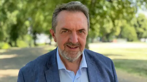 Rob Trigg/BBC Matthew Green, looking directly down the camera. He is standing outside, with grass and trees behind him. Green is wearing a blue linen jacket and an open-necked blue shirt. He has dark hair, flecked with grey and a greying, neatly trimmed beard.