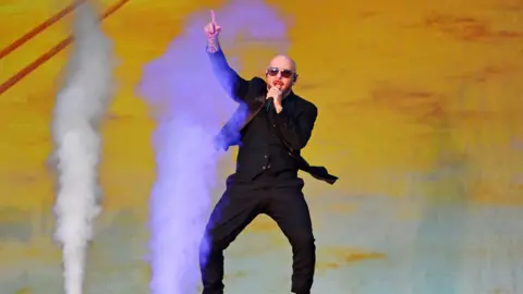 Getty Images Pitbull performs on stage as smoke machines go off in the background. He wears a black suit and sunglasses. 