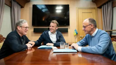 Britain's Prime Minister Keir Starmer wearing a black zip-up jacket sits at a wooden table with French President Emanuel Macron, in white shirt and hoodie, and German Chancellor Friedrich Merz, in blue jumper and shirt, on board a train to the Ukrainian capital Kyiv where all three will hold meetings with Ukrainian President Volodymyr Zelensky
