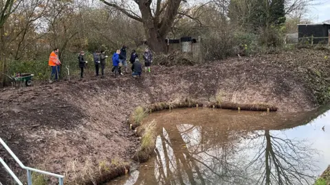 A small water dyke with children walking along the sides to plant shrubs which will act as flood defences