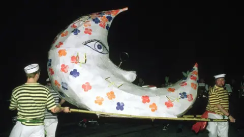 Men dressed as sailors in striped shirts and white trousers hold a large paper moon lantern on a dark night.