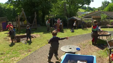 A group of schoolchildren run around a play park with sand pits and climbing frames.