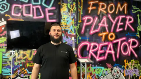 Khaled Ayad, has black hair and beard and wears a black t-shirt. He stands in front of a multi coloured graffiti wall with the name of his business and its motto - from player to creator - sprayed on the wall.