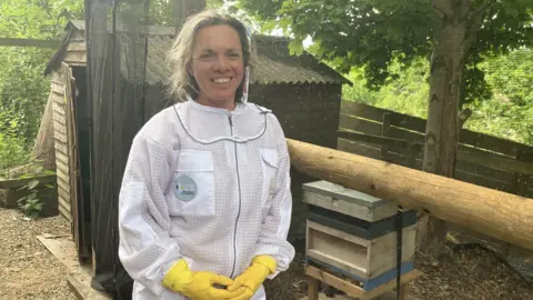 A woman in her early fifties wearing white overalls and yellow rubber gloves. She is smiling and standing against the backdrop of an animal pen, with a wooden hive box and a tree in it.