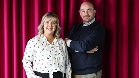 Tara Mills and Declan Harvey standing in front of a red curtain. Tara is wearing a white blouse and Declan has a black jumper on. Both are smiling towards the camera.