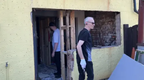 A man with white hair, black clothing and sunglasses walks past the vandalised building