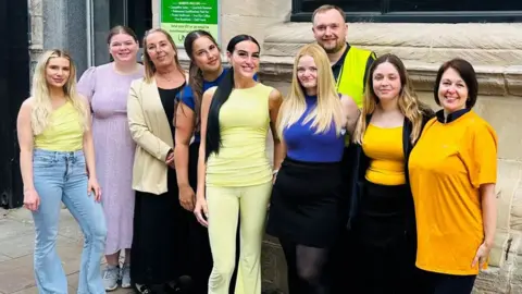 Stand Up For Southport Nine people stood in a row outside a stone building front. They are wearing a mix of wearing yellow outfits in a variety of shades.