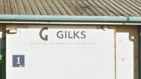 A sign that says "Gilks" on the side of a white building with a roof above